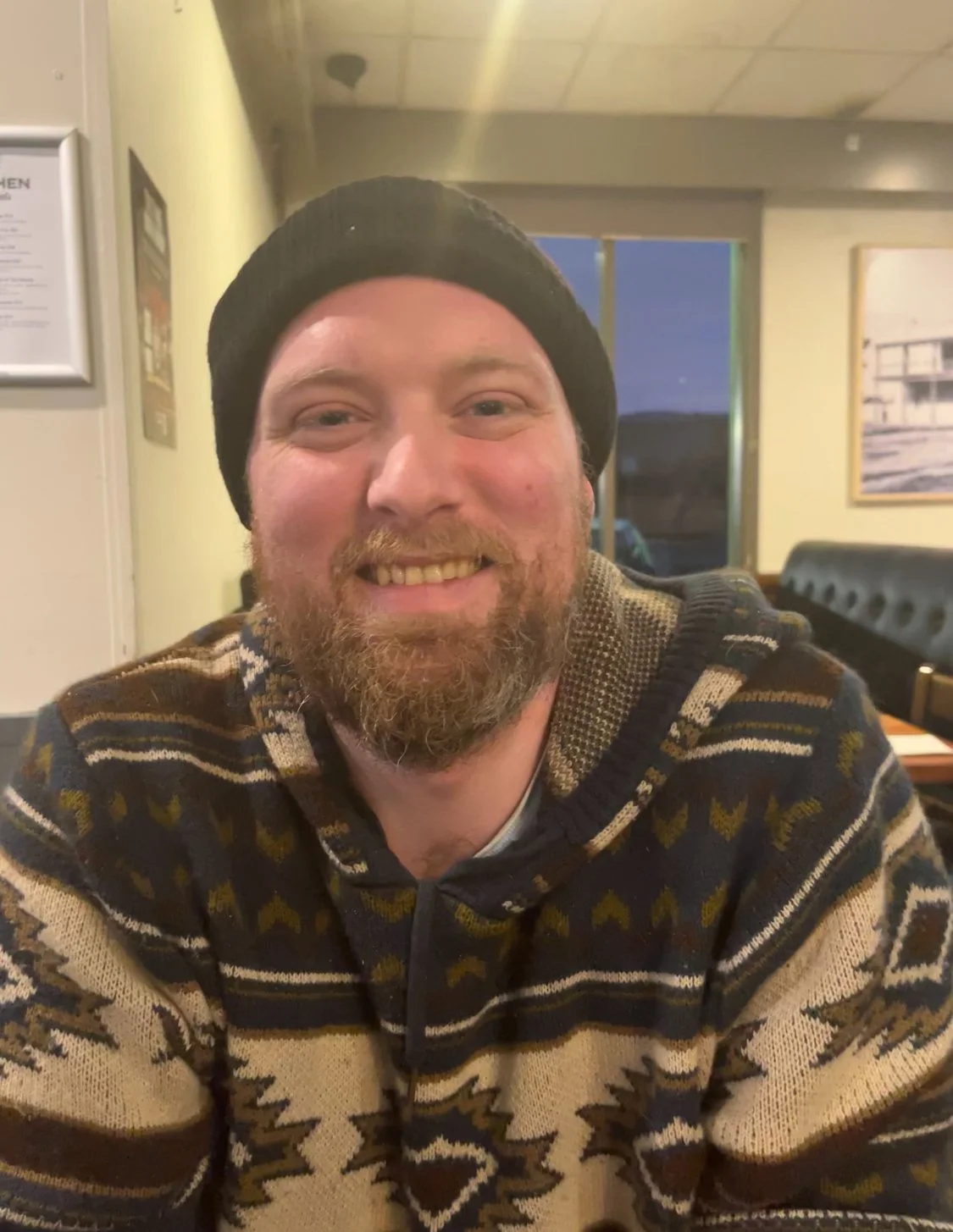 Man smiling with a beard, wearing a black beanie and a patterned sweater, sitting inside a restaurant.