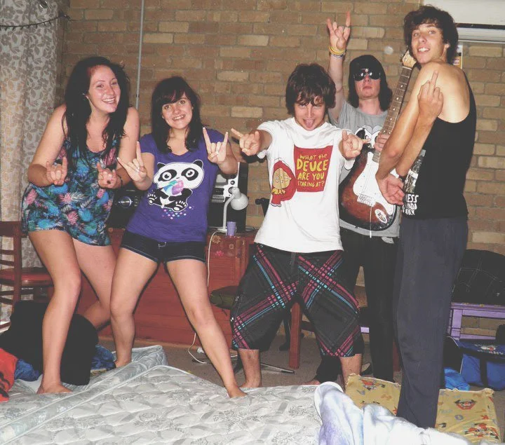Five young people posing in a room with a brick wall, some making hand gestures and smiling, with a guitar and other items in the background.