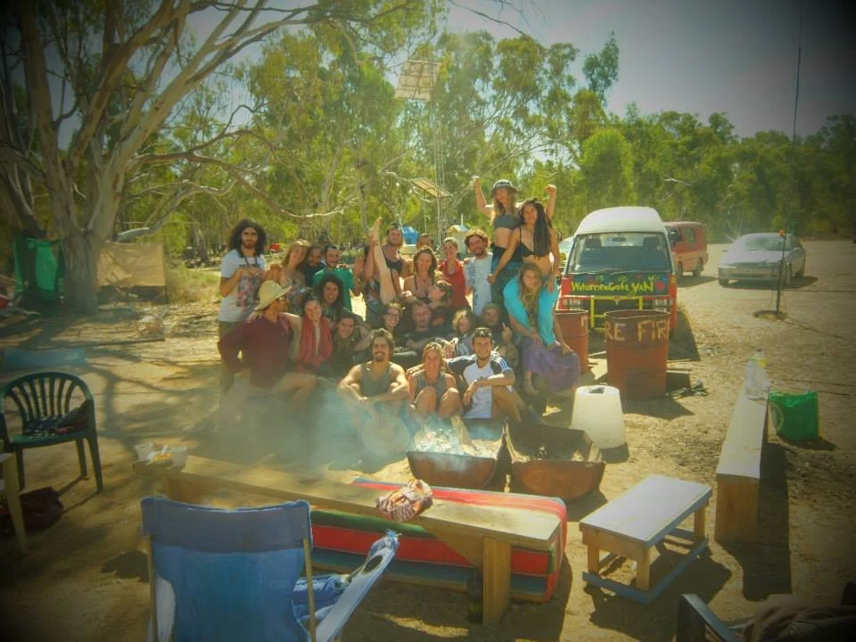 A large group of people gathered outdoors around a campfire with trees and vehicles in the background, some standing, sitting, and posing for the photo.