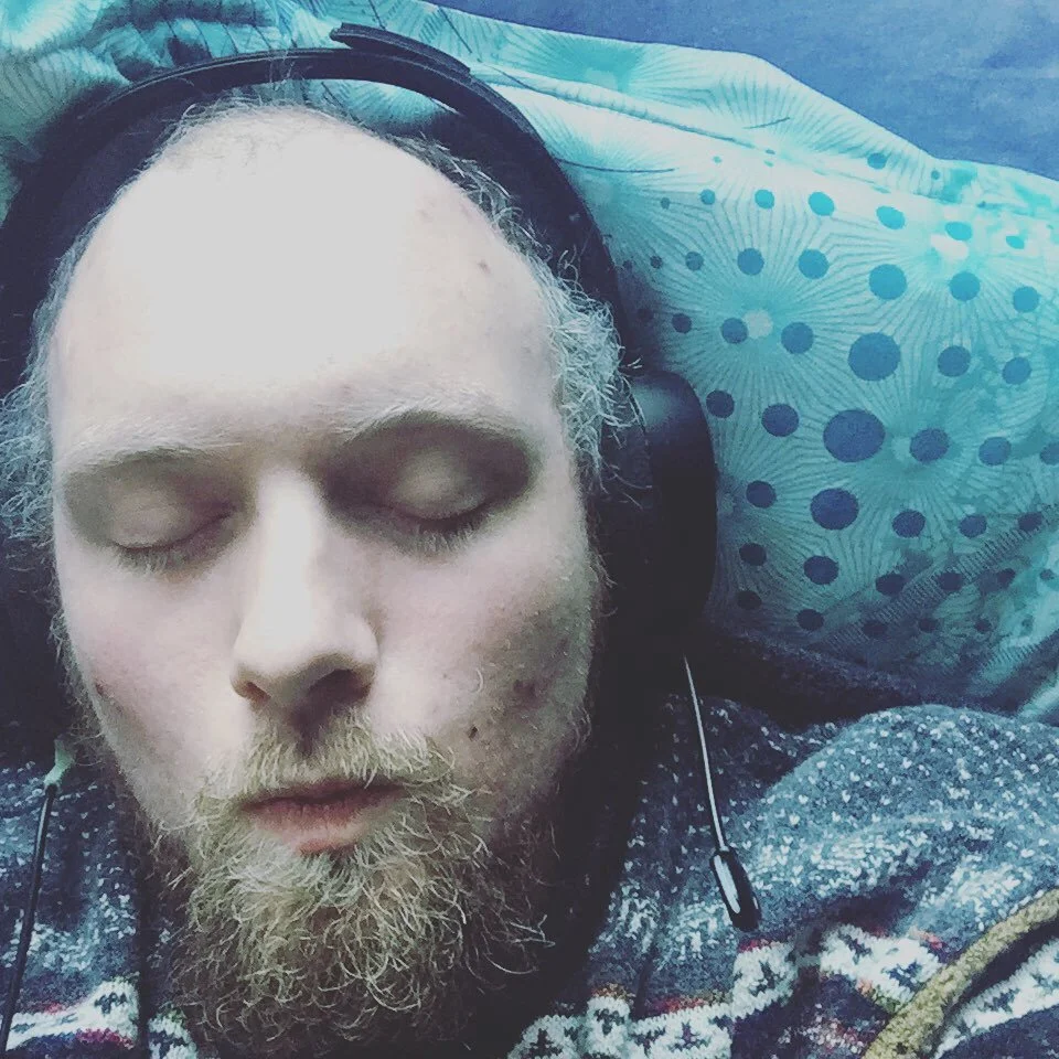 A man with a beard and closed eyes, wearing headphones, resting on a pillow with a blue and green dotted pattern.