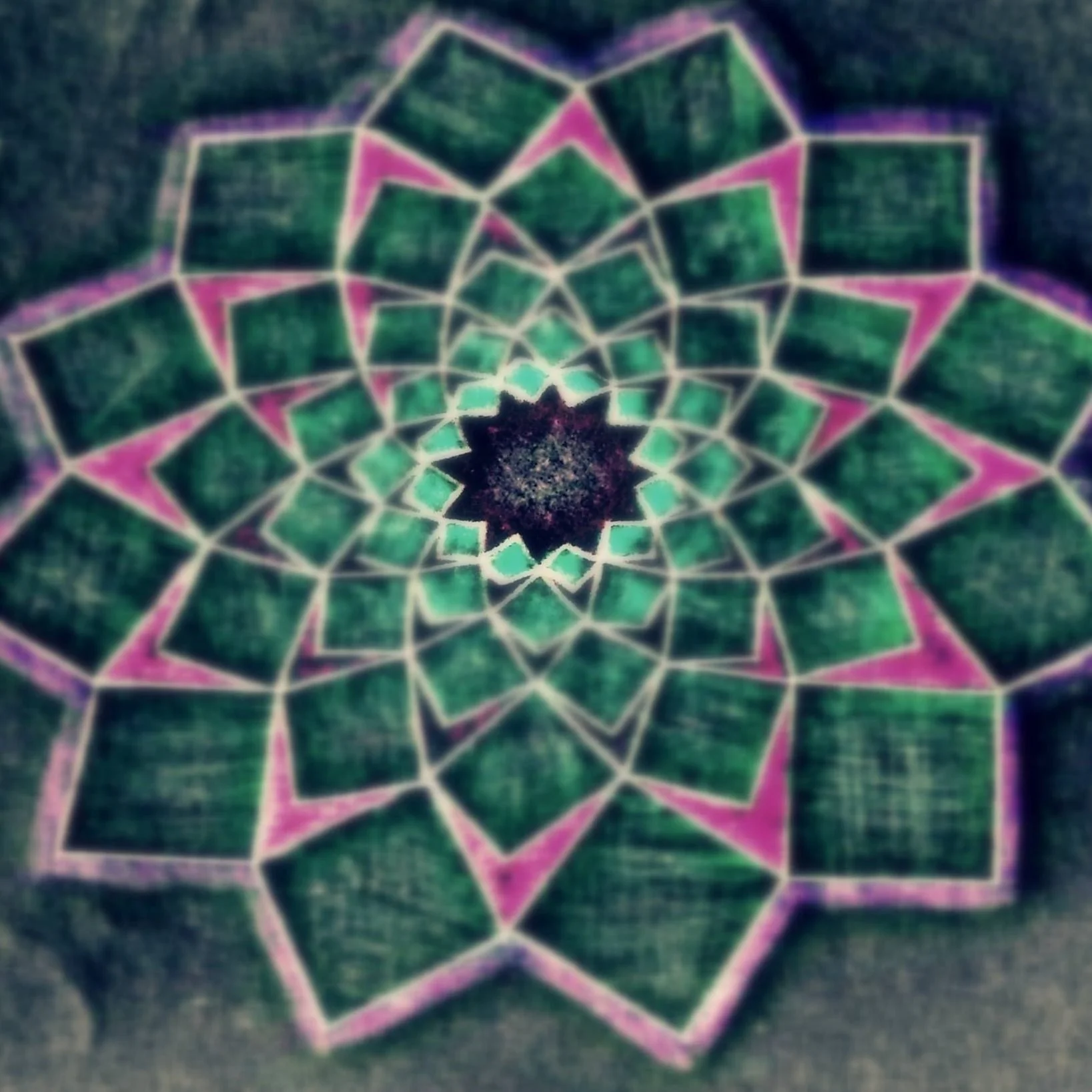 A colorful chalk drawing of a mandala with geometric patterns and a star shape at the center, featuring green, pink, and purple colors.