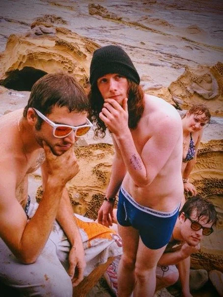Four young men at the beach with sandy rocks in the background, some wearing sunglasses and one in a black beanie, one shirtless.