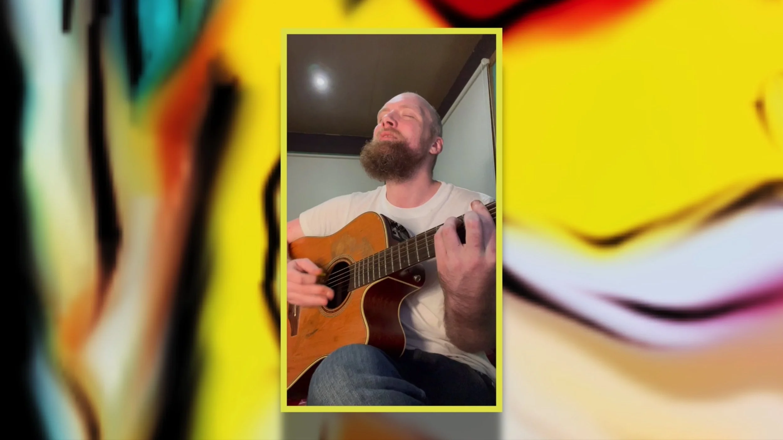 A man with a beard and closed eyes is playing an acoustic guitar with a white shirt. He appears to be singing or enjoying the music in a room with a plain background.