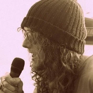 Profile of a person wearing a knit beanie, holding a microphone, with long curly hair.