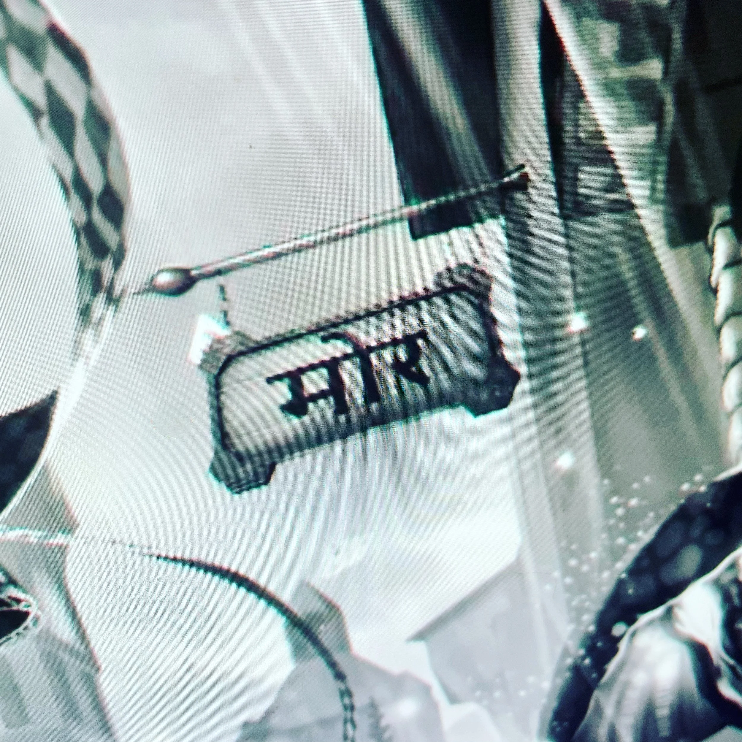 Close-up of a street sign with the word 'Mare' in Hindi script, mounted on a pole, with a background of tall buildings and overcast sky.