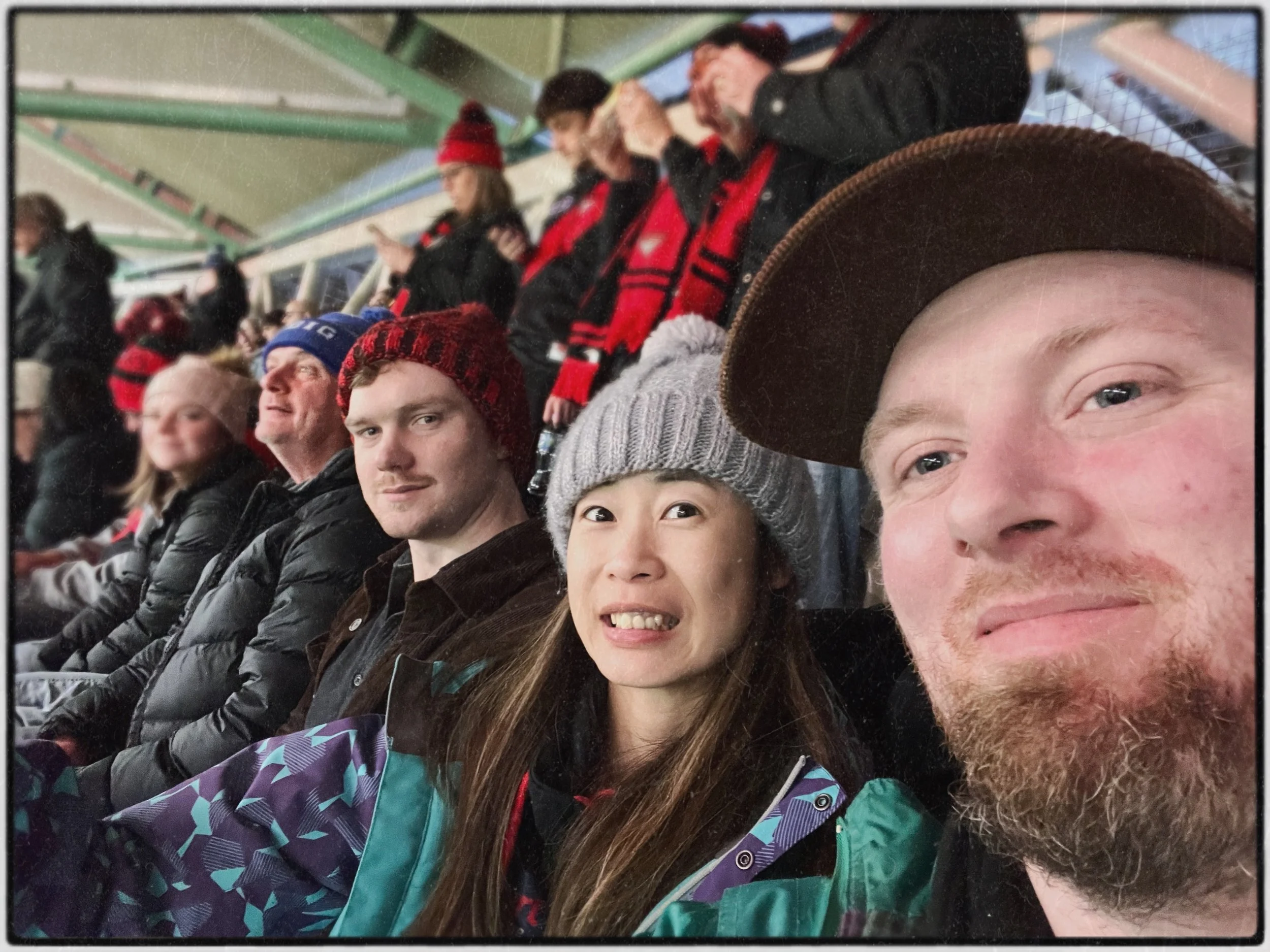 A group of people sitting in stadium seats at an outdoor event, wearing winter clothing including hats and jackets, with some standing in the background.