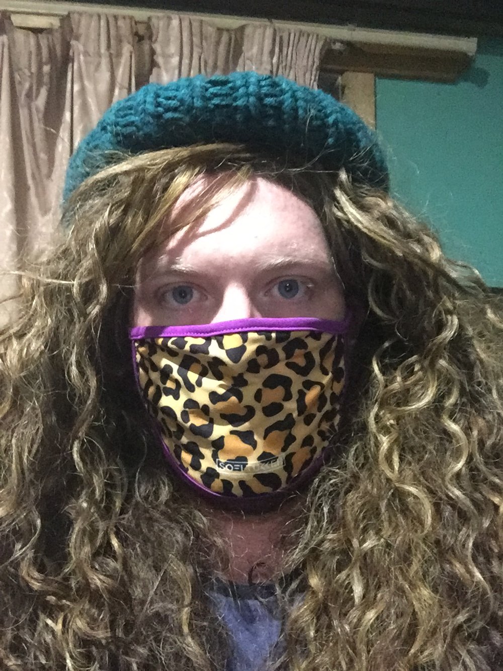 Person with long curly hair wearing a blue knit hat and a leopard print face mask, indoors with beige curtains and a green wall in the background.