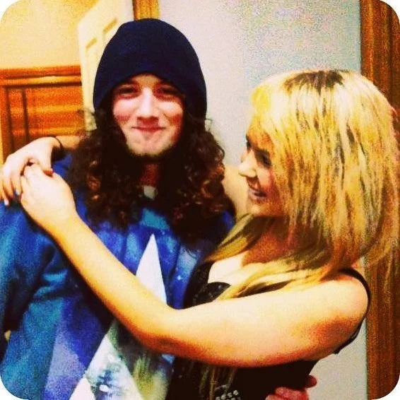 A young man with long curly hair, wearing a black beanie and a blue hoodie, smiling while embracing a young woman with long blond hair with dark streaks, wearing a black top, and smiling back.