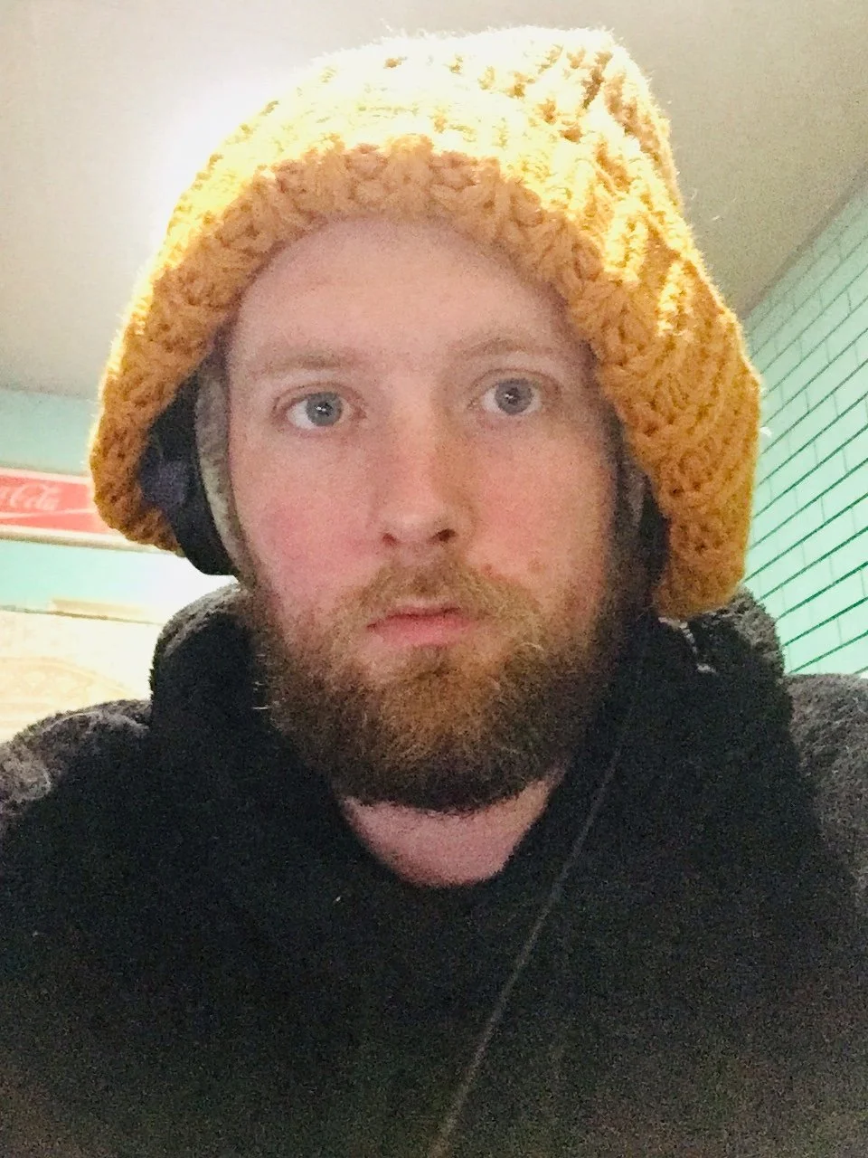 A man with a beard and light blue eyes wearing a yellow knitted hat and black hoodie indoors.