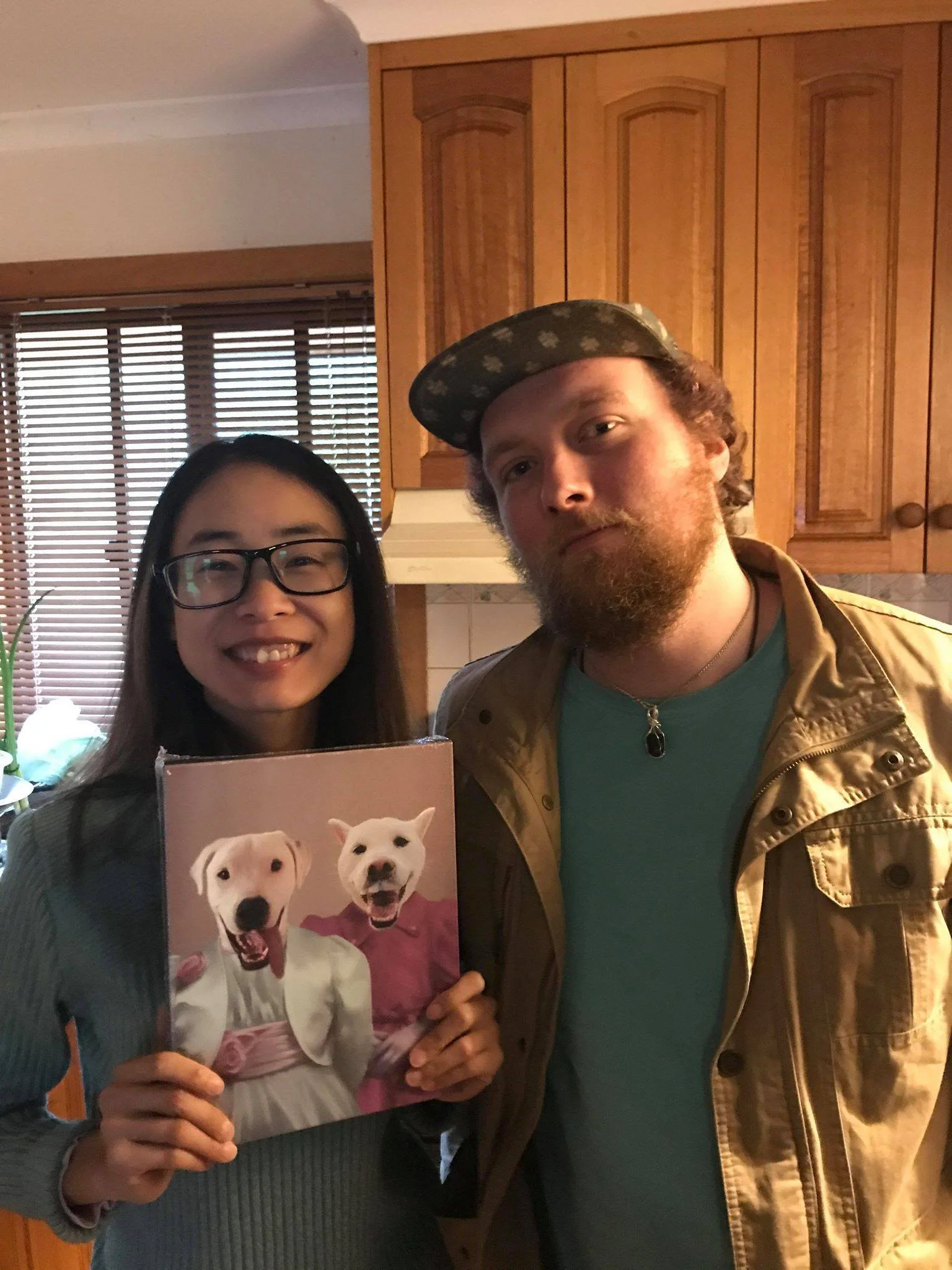 A woman smiling and holding a picture of two dogs dressed as humans, standing next to a man in a kitchen with wooden cabinets and blinds on the window.