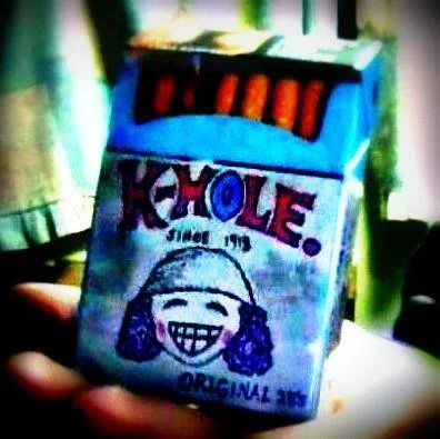 Colorful packet labeled 'K-HOLE' with a cartoon character smiling and wearing headphones. Original Cigarette pack design by Cale Anderson.