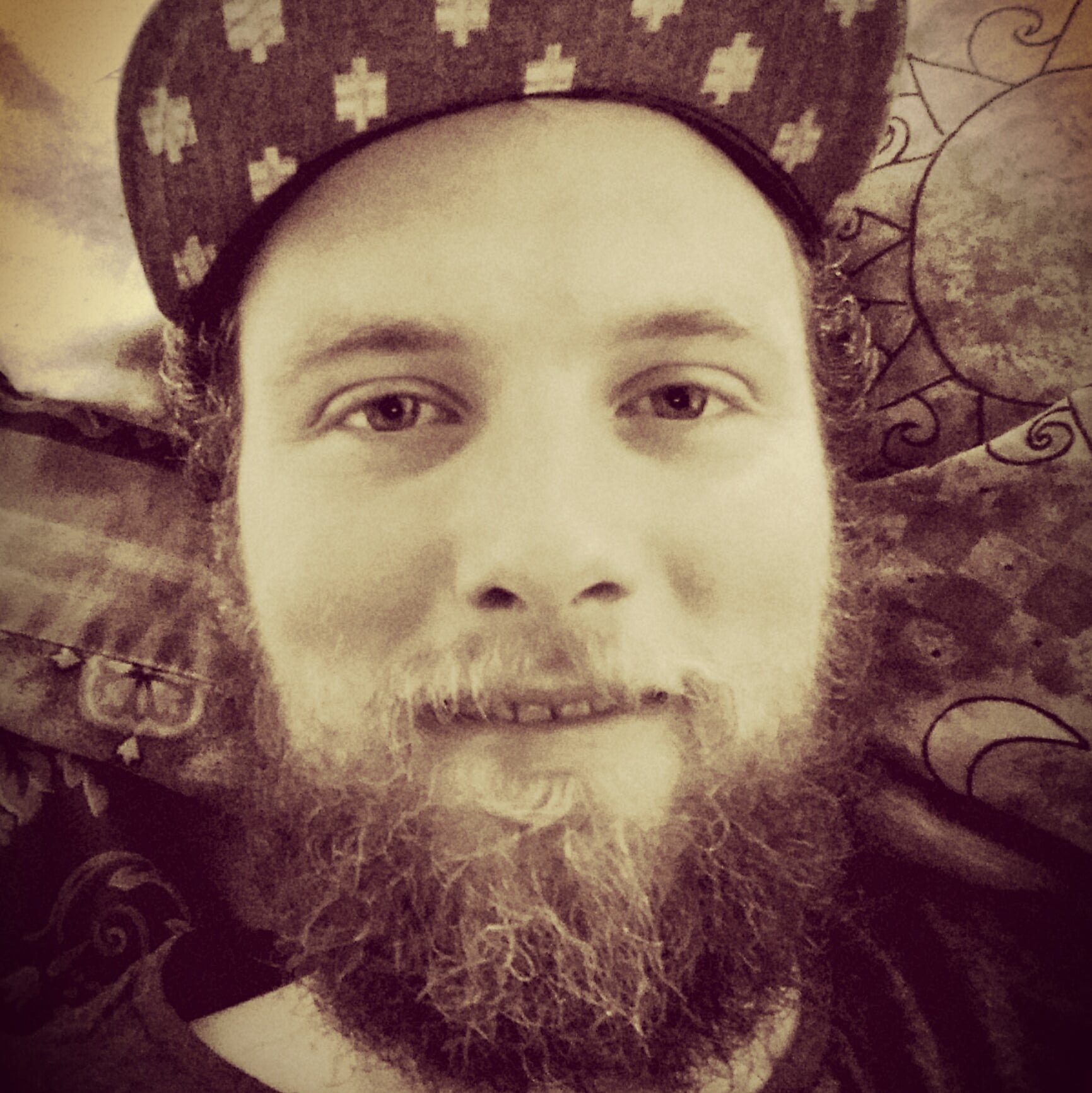 A person with a full beard and mustache, wearing a patterned cap, lying on a bed with decorative pillows.