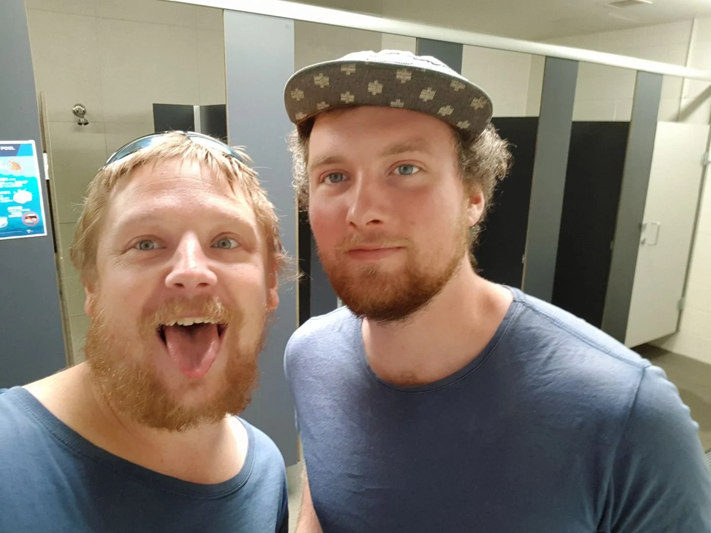 Two young men with beards and casual clothing taking a selfie in a locker room, with lockers and a poster visible in the background.