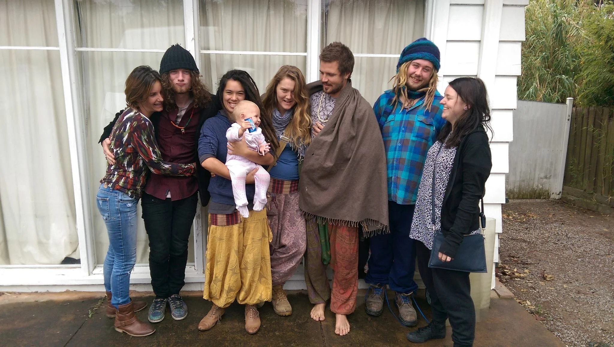 Group of people standing outdoors in front of a white building, smiling and laughing, including adults and a child in colorful clothing, some dressed in casual and others in bohemian style, with a mix of barefoot and shoe-wearing individuals.