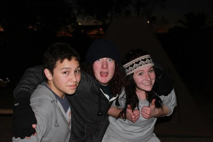 Three teenagers outdoors at night, smiling, with two giving thumbs up, in front of a dark tent or teepee.
