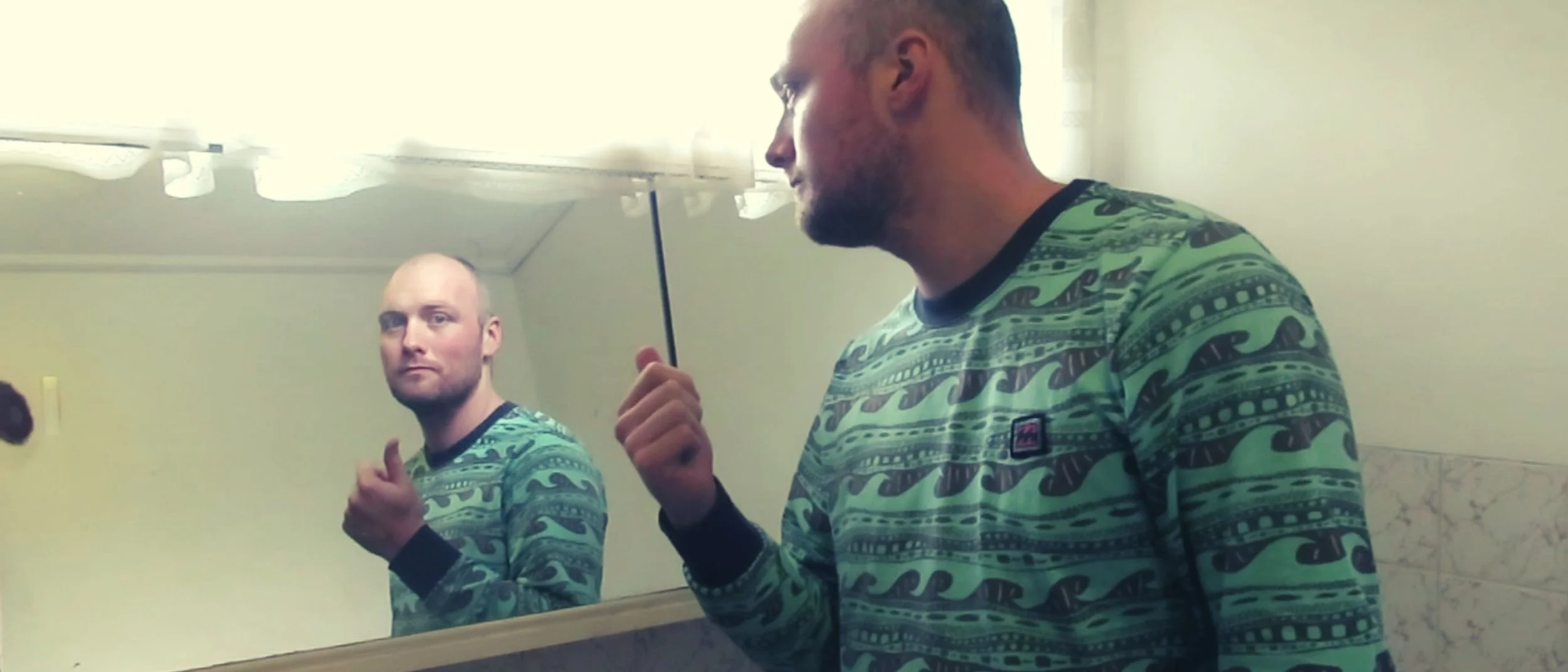 Two men in patterned green shirts standing in front of a mirror, one looking at the camera and the other looking at his reflection, in a bathroom with a light fixture and marble-tiled wall.