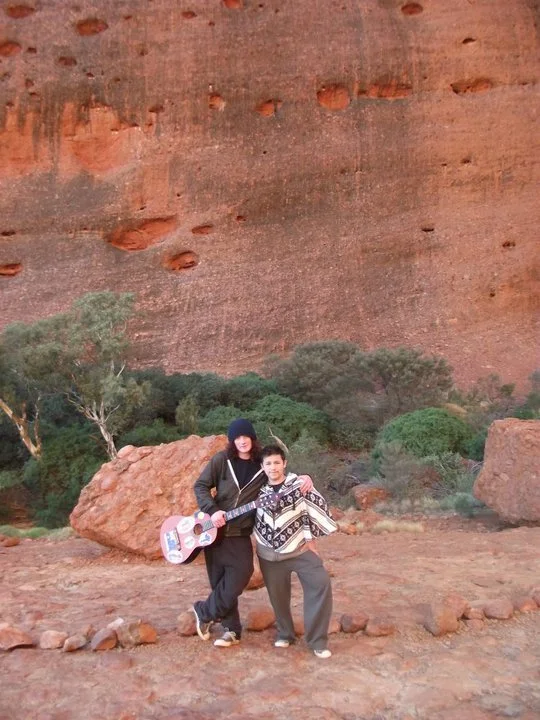 Two people standing outdoors on rocky terrain, one holding a pink guitar, with a large reddish rock formation and green bushes in the background.