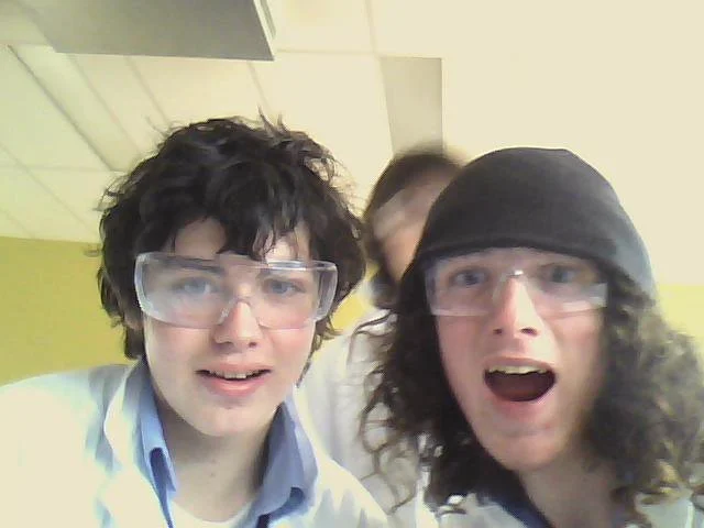 Two young men wearing safety goggles, one with curly dark hair and a light blue shirt, and the other with long curly hair, a black hat, and a white shirt, in a laboratory or classroom setting.