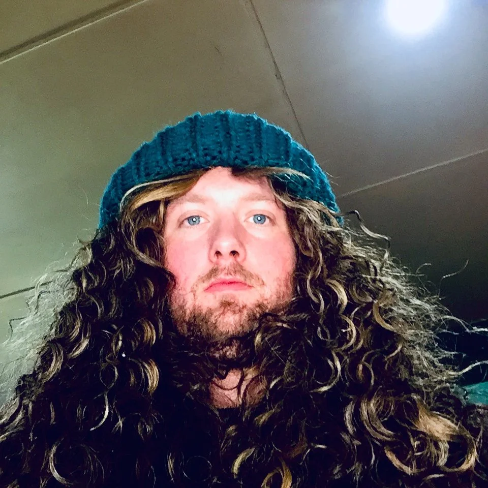 A person with long curly hair wearing a blue knitted beanie, looking at the camera with a neutral expression, standing indoors under a ceiling light.
