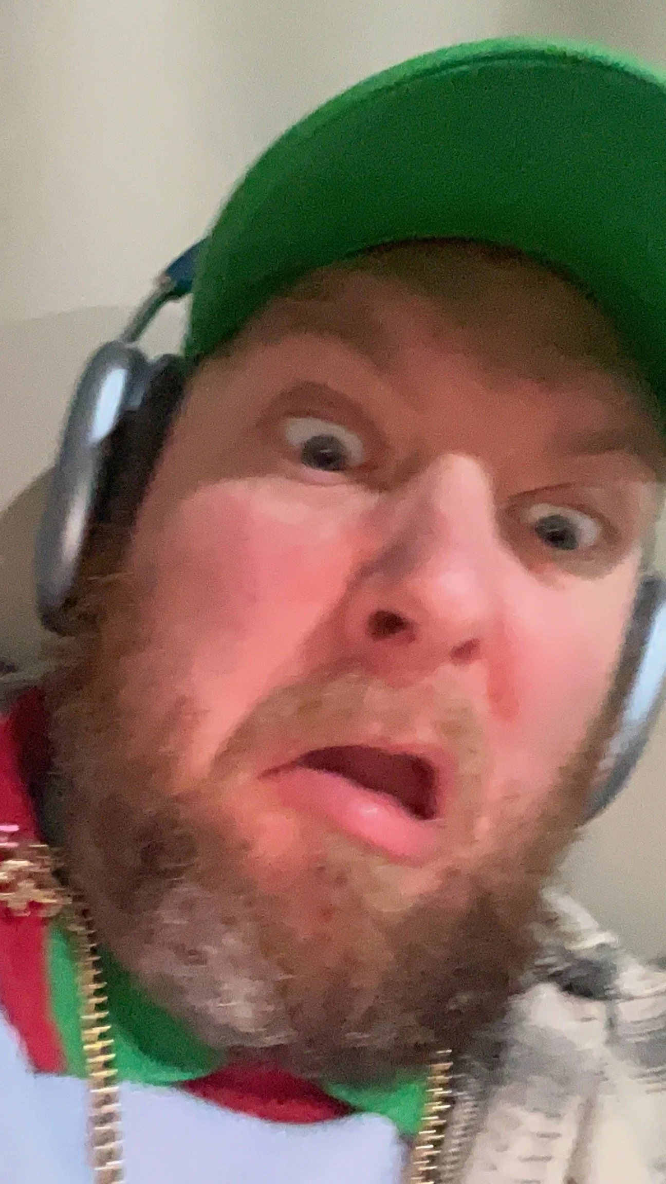 A man wearing a green baseball cap, headphones, and a jacket with colorful stripes, making a surprised or confused facial expression.