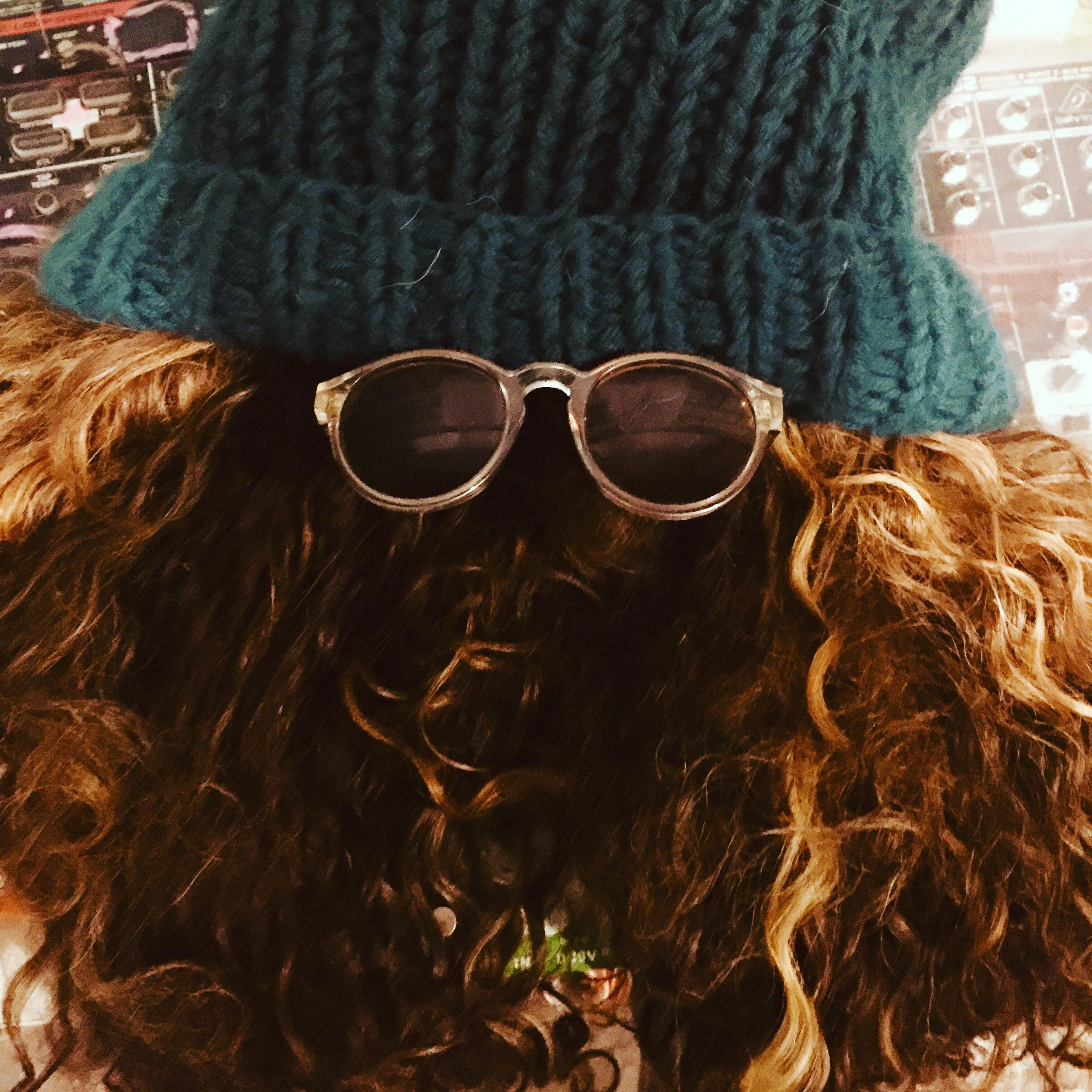 A top-down view of a person with long, curly brown hair wearing round sunglasses and a knitted dark teal hat, with a background of various small items.