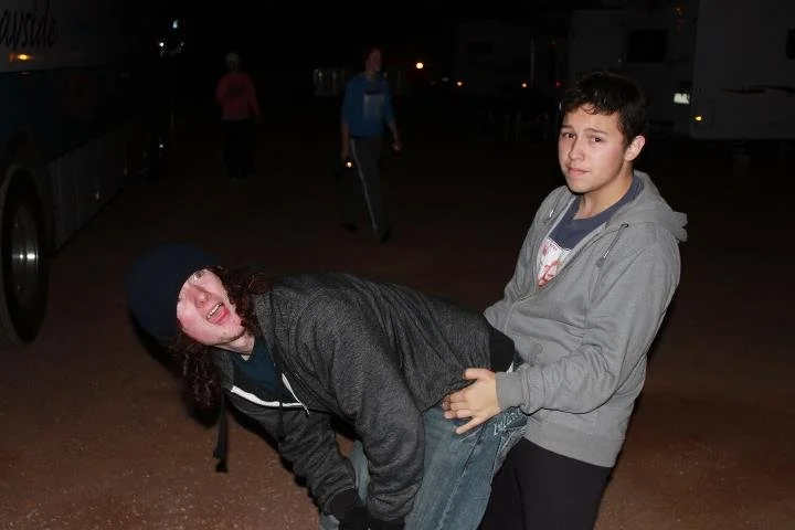 Two young people, one standing and holding the other, who appears to be pretending to be in pain, outdoors at night with other people in the background.
