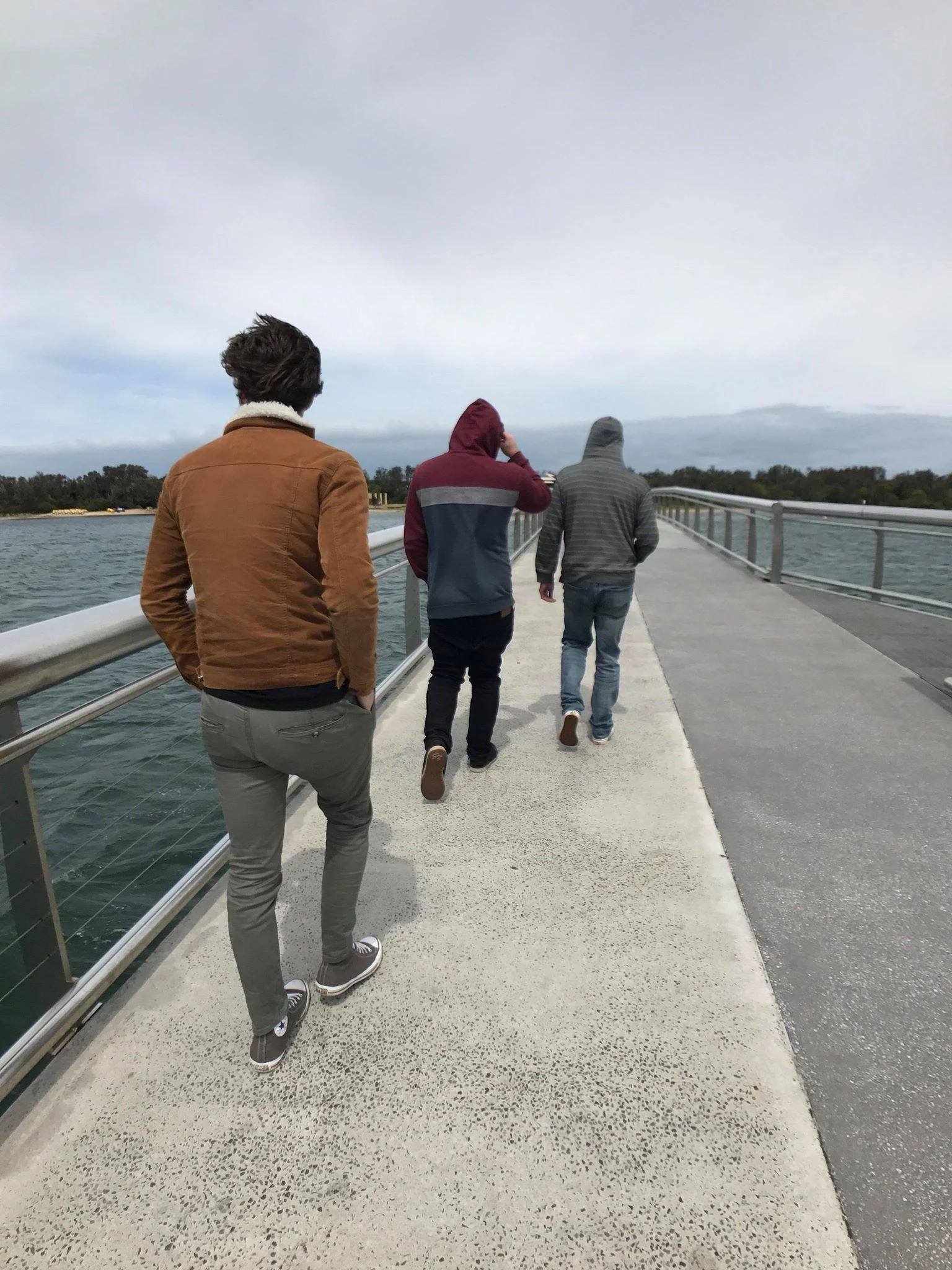 Four young men walk along a pier over water, wearing hoodies and jackets, with a cloudy sky overhead.