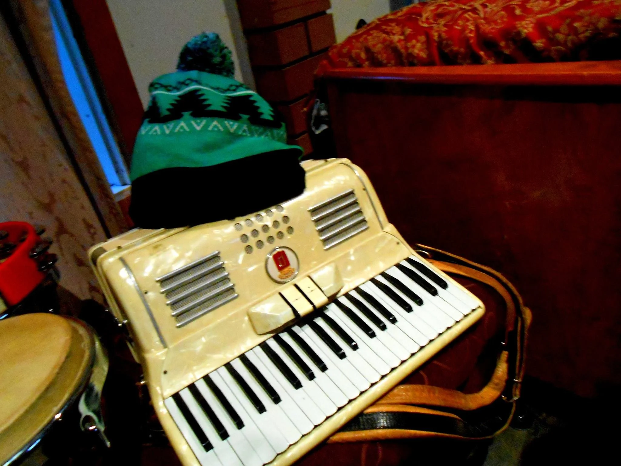 A vintage beige toy keyboard with black and white keys, a blue and green knitted beanie with a pom-pom on top placed on it, sitting on a wooden surface in a cozy room.
