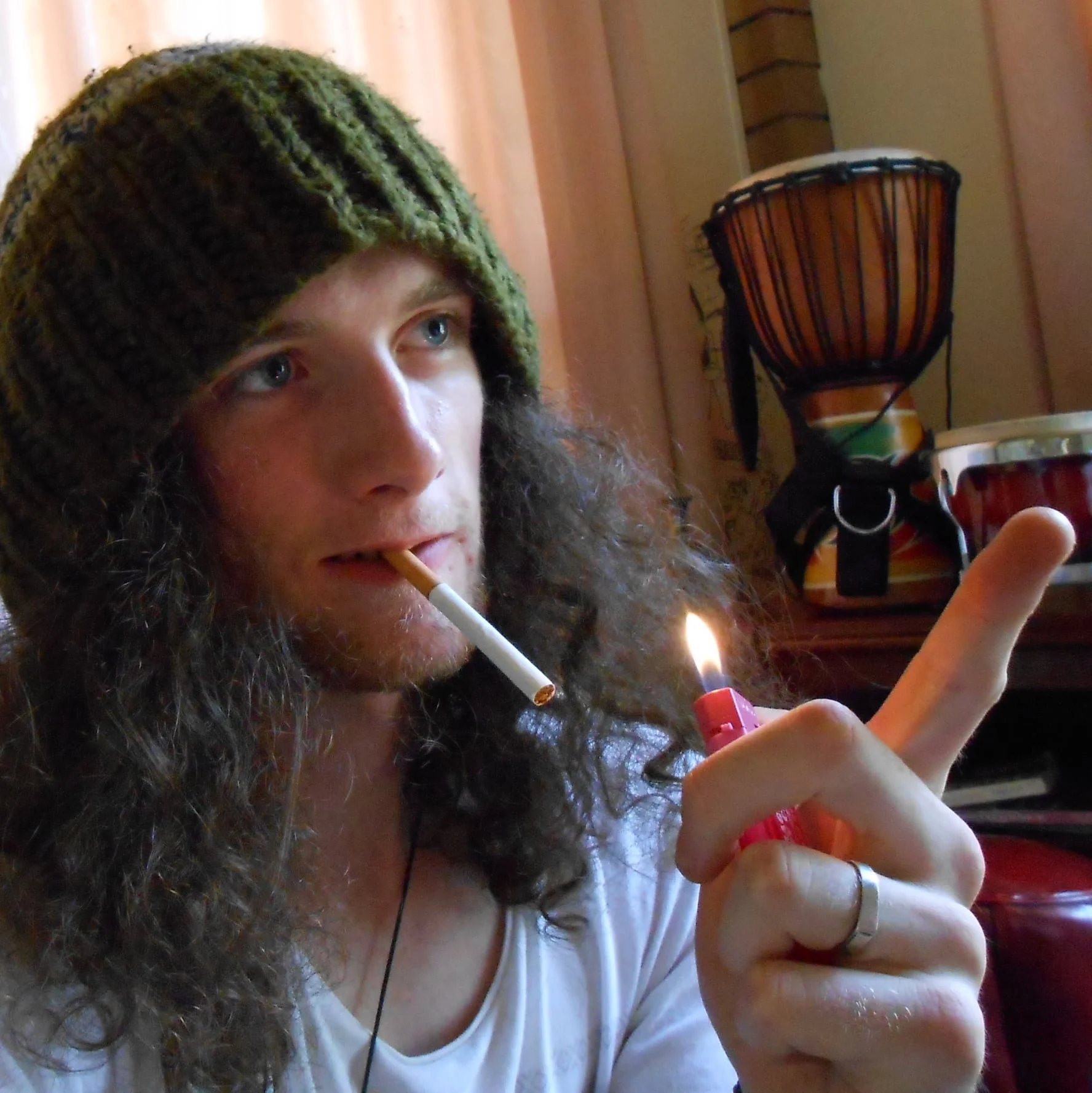 A person with long curly hair, wearing a knit hat, is lighting a cigarette with a lighter that has a small flame. The person is pointing upward with their index finger. In the background, there is a drum and some household items.