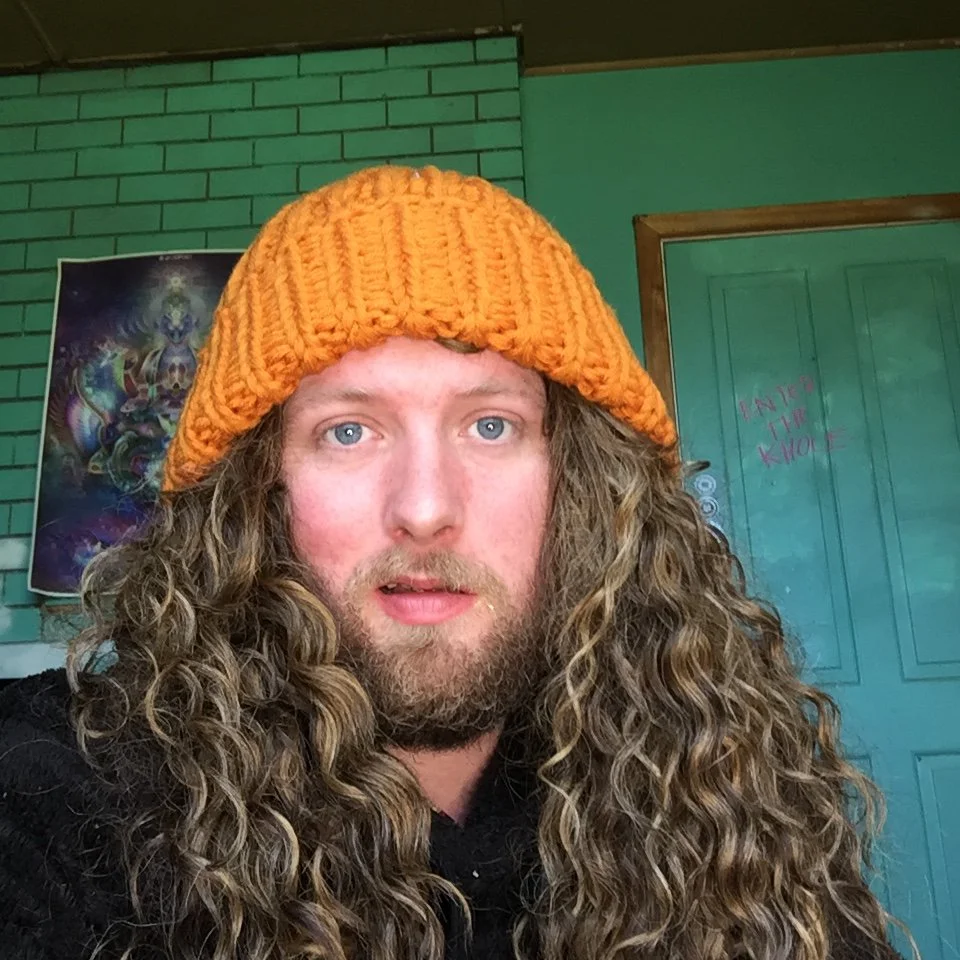A man with long, curly hair and a beard wearing an orange knit beanie.