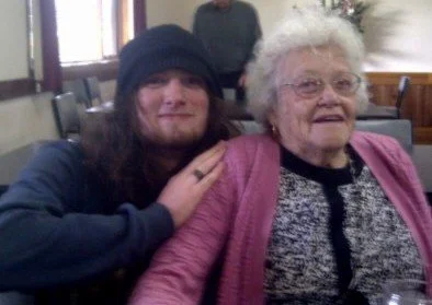 A young man with long dark hair and a black beanie sitting next to an elderly woman with curly white hair, glasses, and a pink sweater, both smiling in a room with chairs and windows in the background.