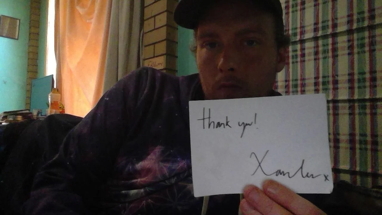 A man wearing a dark cap and shirt is sitting indoors, holding a white sign that says 'thank you! XamLxx' in front of a colorful plaid curtain.