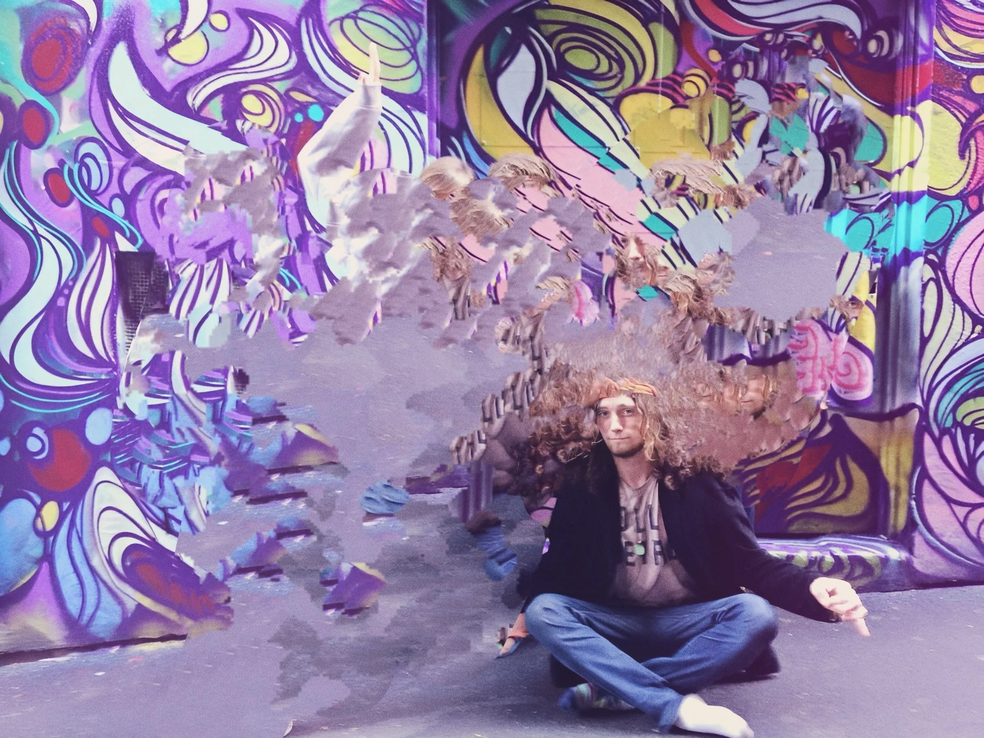 A man with long, curly hair sitting cross-legged on the floor in front of a colorful, abstract graffiti mural, with digital distortion effects.