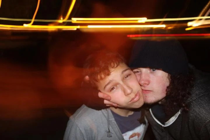 Two young people taking a close-up selfie at night with streaks of street lights in the background.