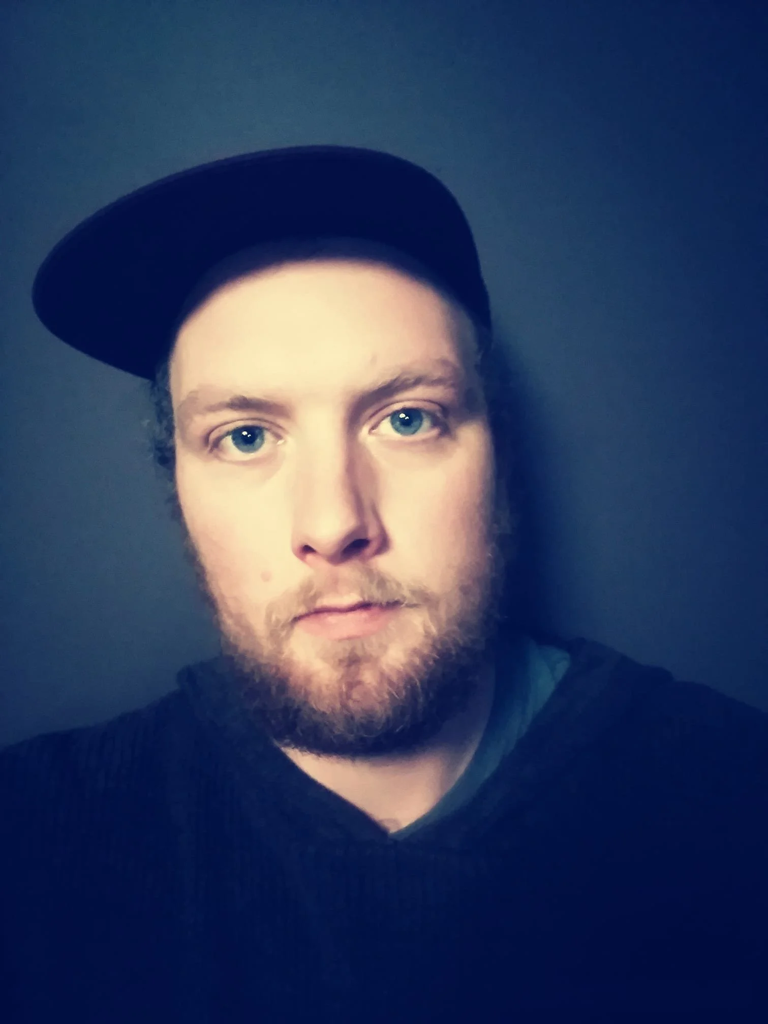 A young man with light skin, blue eyes, and a beard, wearing a black baseball cap tilted to the side, against a dark background.