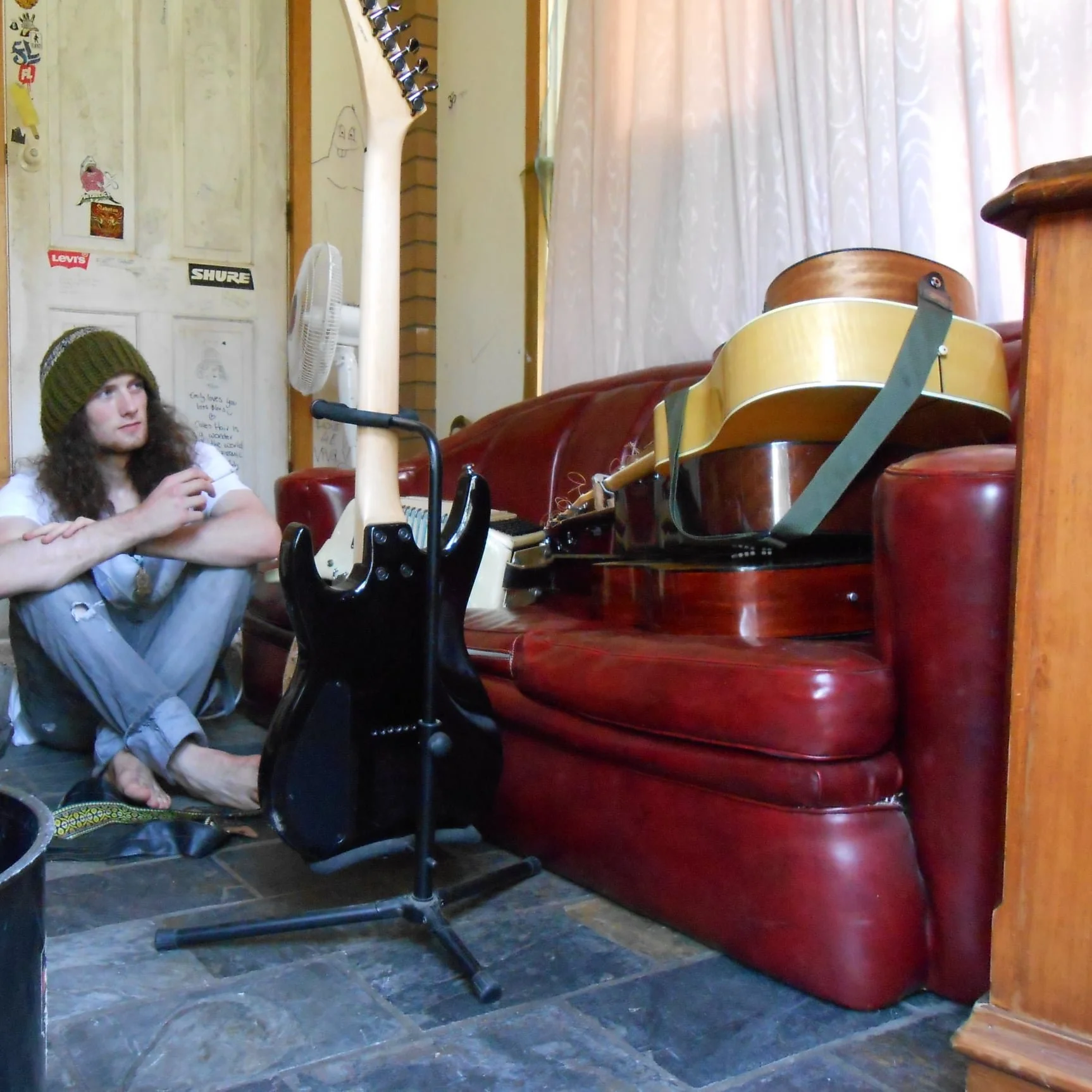 Young person with long hair wearing a green knit hat and ripped jeans sitting on a dark tiled floor, looking at a collection of guitars and musical equipment against a red leather sofa in a cozy room.