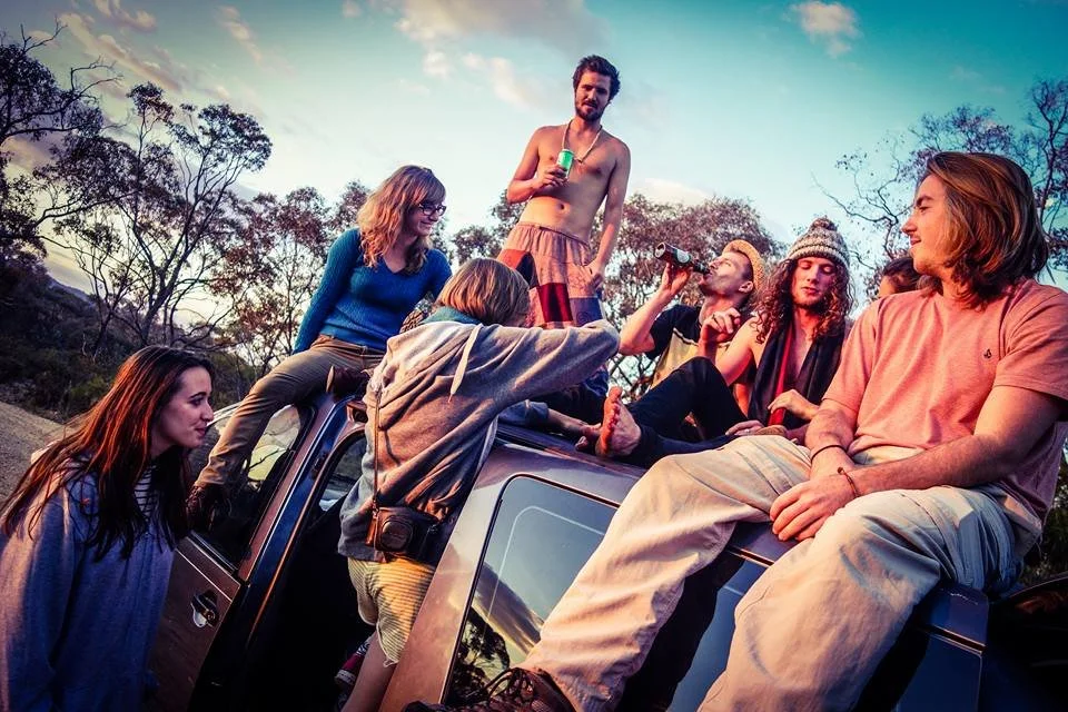 Group of friends hanging out outdoors on top of a vehicle during sunset, some sitting, others standing and drinking, with trees and a colorful sky in the background.