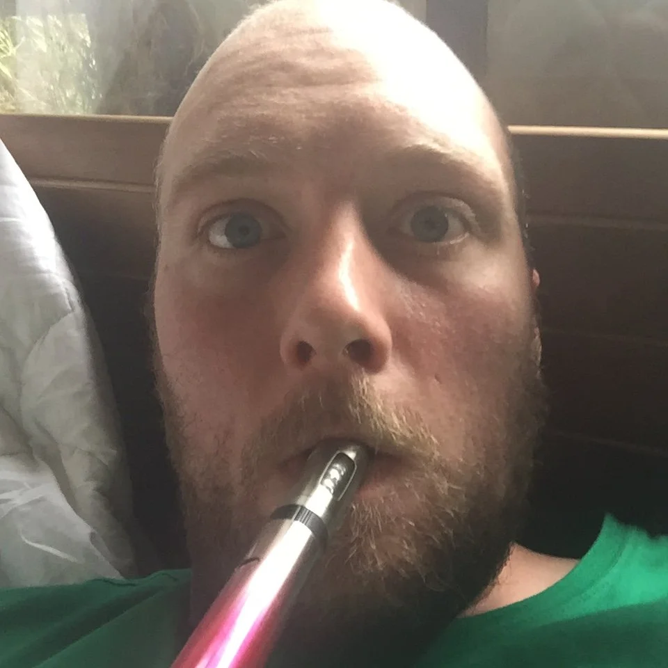 A man with a beard and blue eyes lying in bed, holding an electronic cigarette in his mouth.