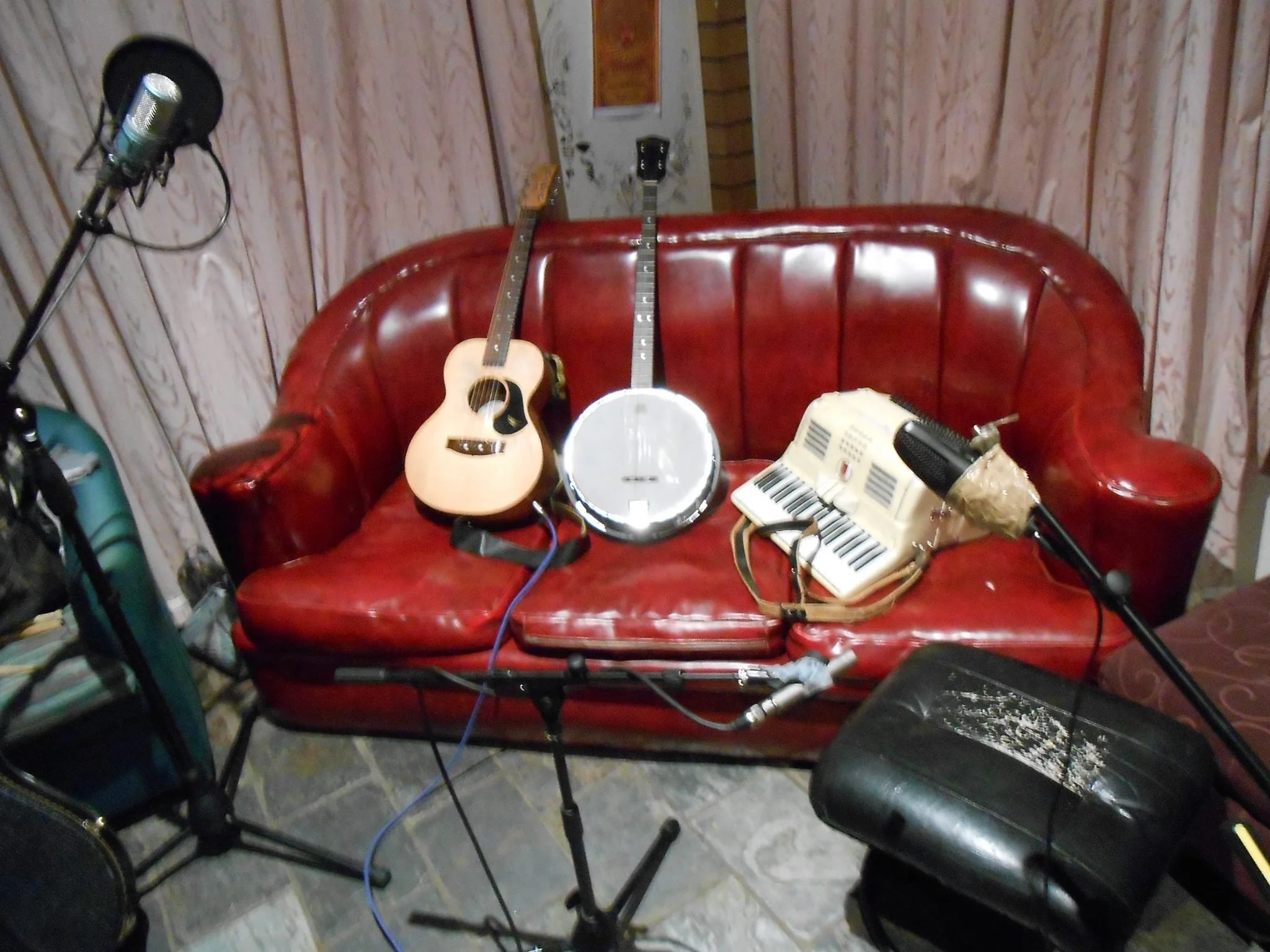 A vintage music studio setup on a red leather sofa with an acoustic guitar, a banjo, a toy keyboard, and a microphone stand, in a room with curtains and a tiled floor.