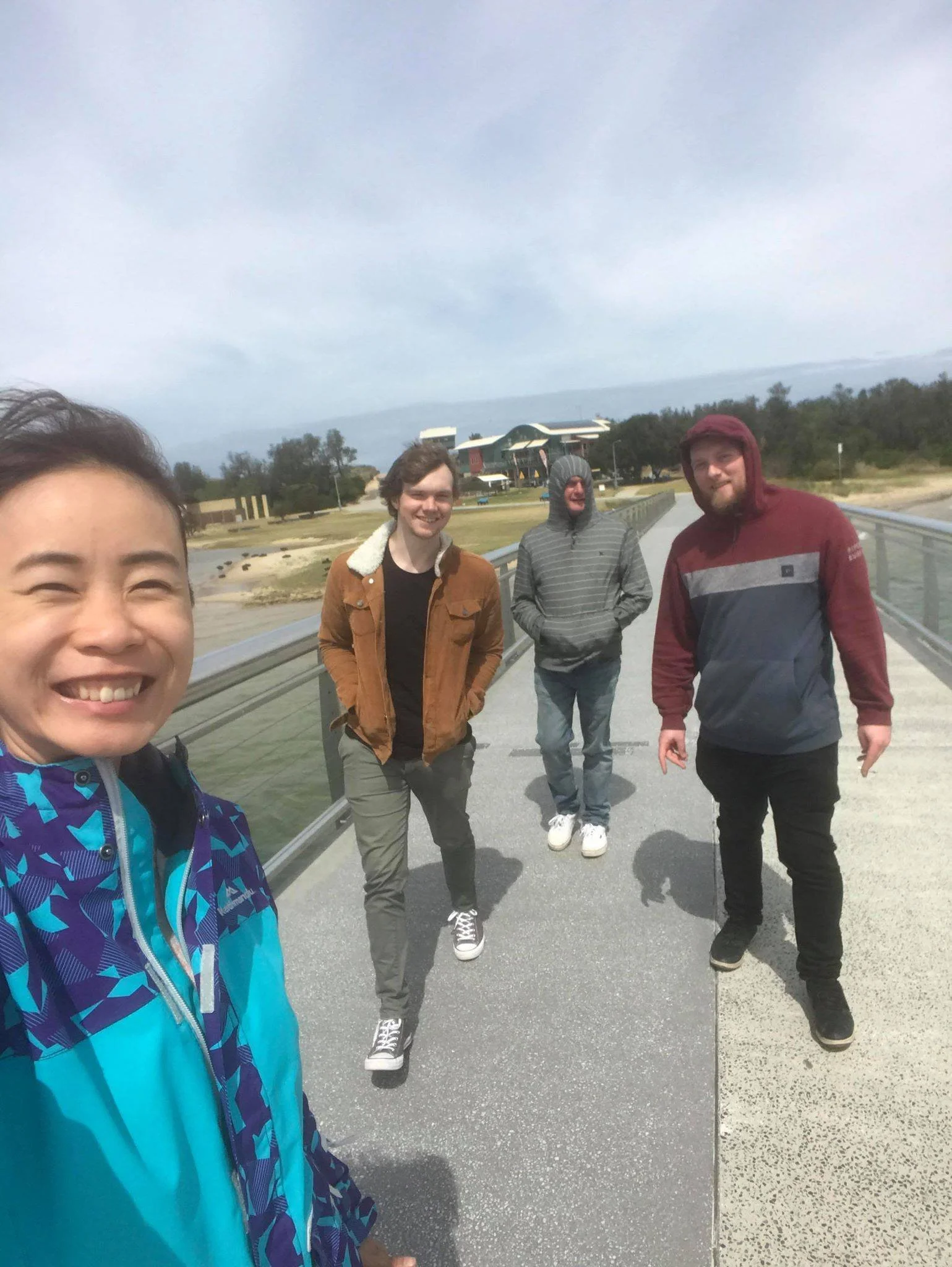 A group of four young adults taking a selfie on a bridge outdoors with a wetland and modern buildings in the background, a partly cloudy sky above them.
