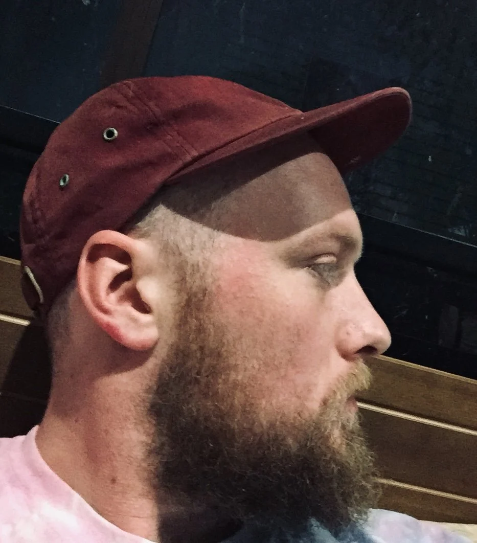 Profile of a man wearing a red cap, with a phased haircut and beard, against dark and wooden background.