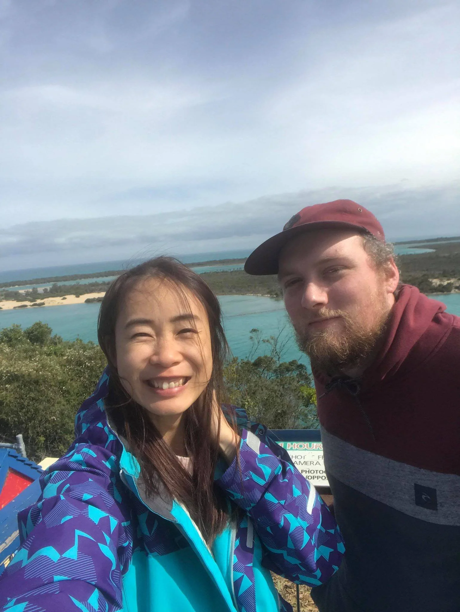 A smiling woman and a man taking a selfie outdoors with a scenic view of a body of water, sand dunes, and trees in the background.