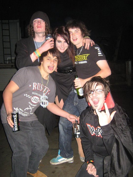 Group of five young people at a party, smiling and making fun poses, holding drinks and cans.