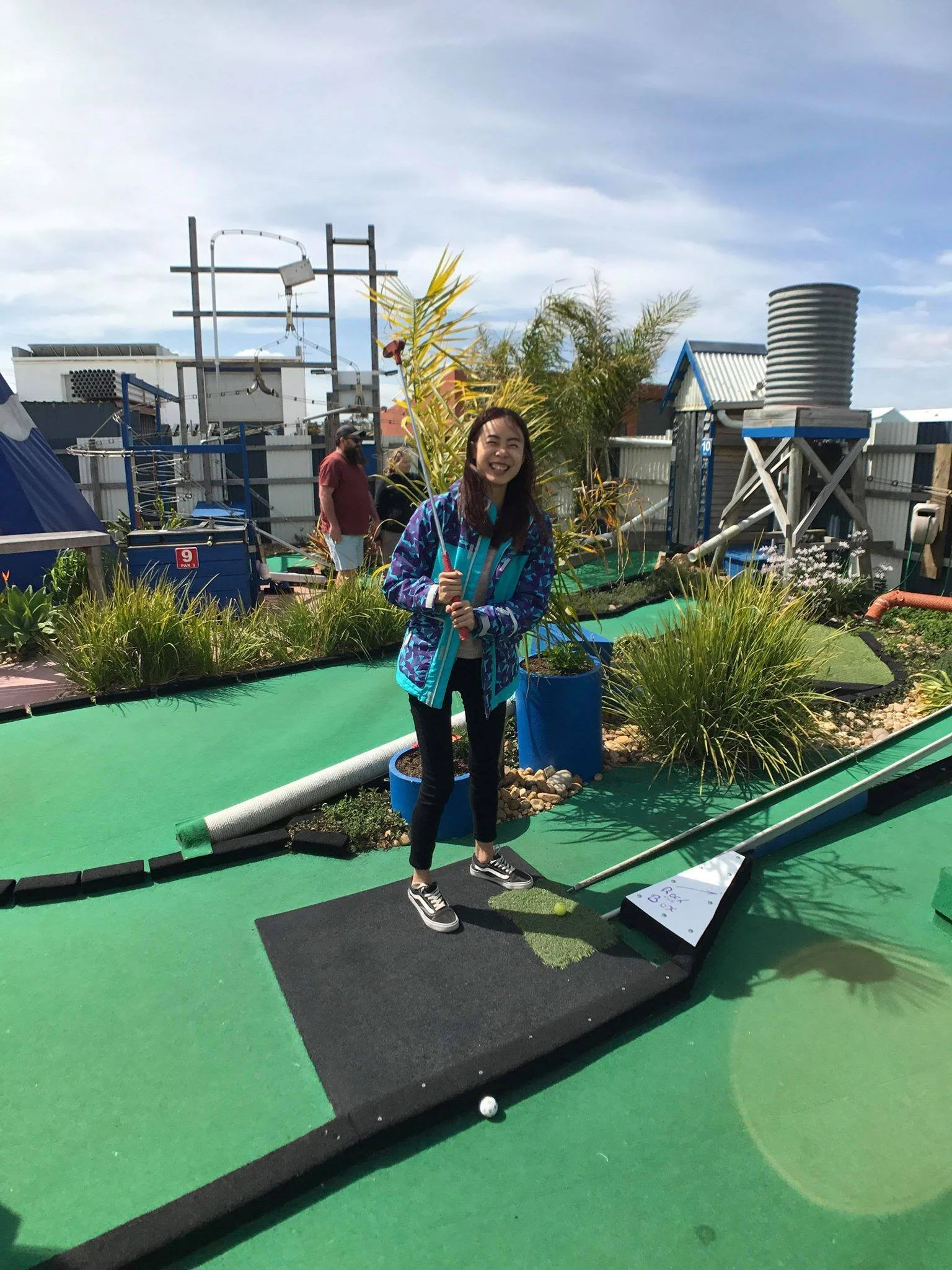 A woman holding a mini golf club and smiling at a mini golf course. The course features artificial turf with a ball and a hole. There are plants and structures in the background under a partly cloudy sky.