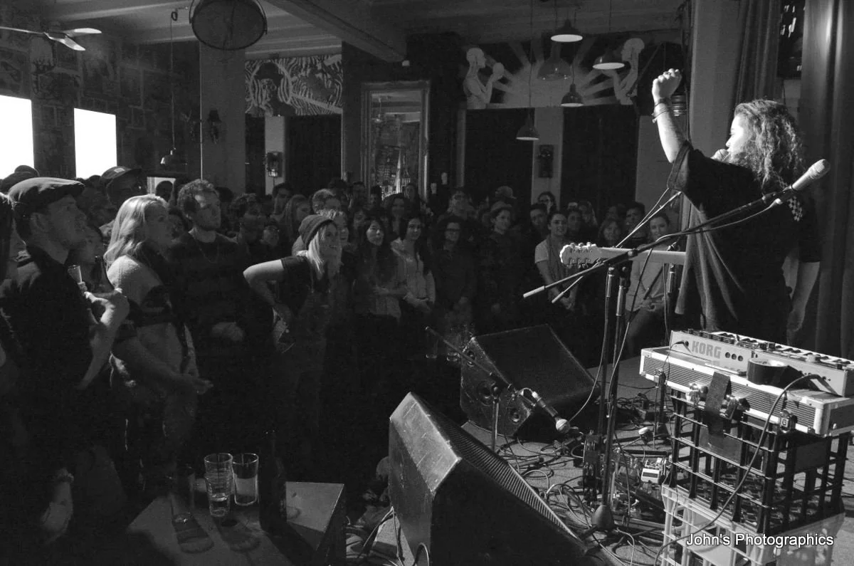 Female singer performing on stage with guitar, microphone, and keyboard in front, audience watching in a bar or club setting, black and white photo.