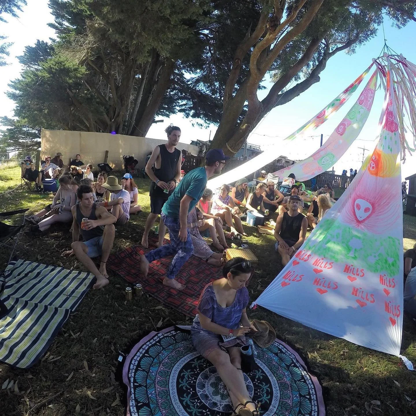 Group of people outdoors at a festival or gathering, sitting on blankets and chairs under a large tree, with colorful decorations and tents.