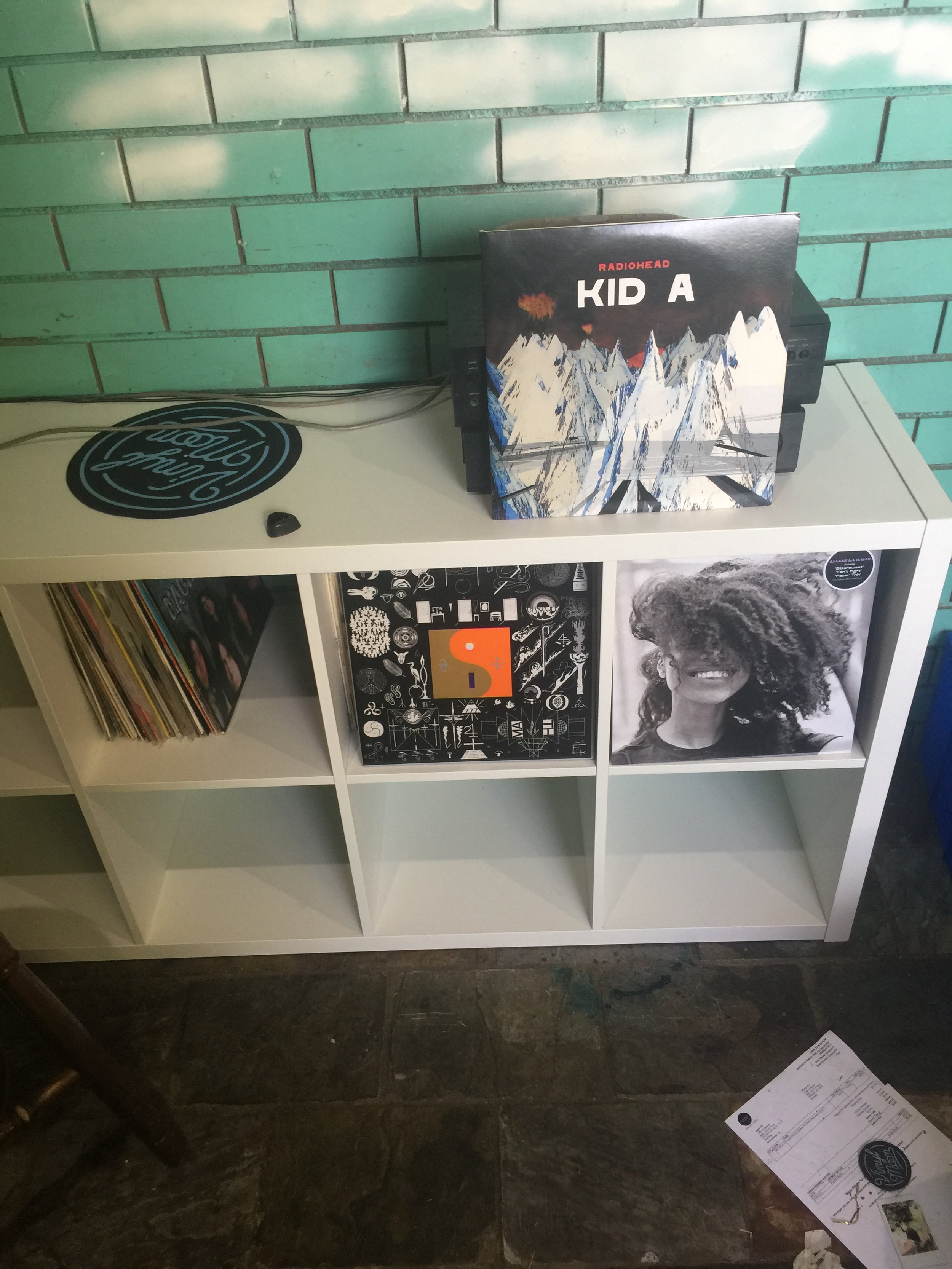 White cubical shelf with vinyl records, a poster of a black and white woman with curly hair, and graphic artwork, against a teal brick wall.