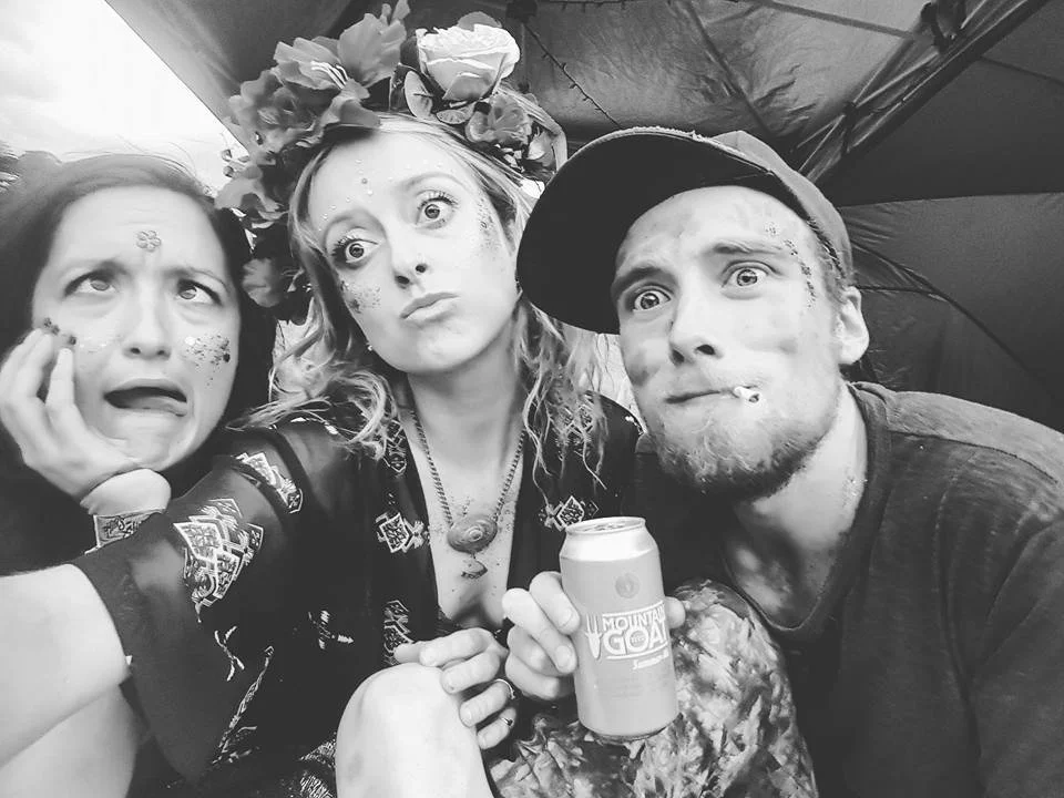 Three people making silly faces, one woman wearing a flower crown, another with face paint, and a man wearing a baseball cap, sitting under a tent.