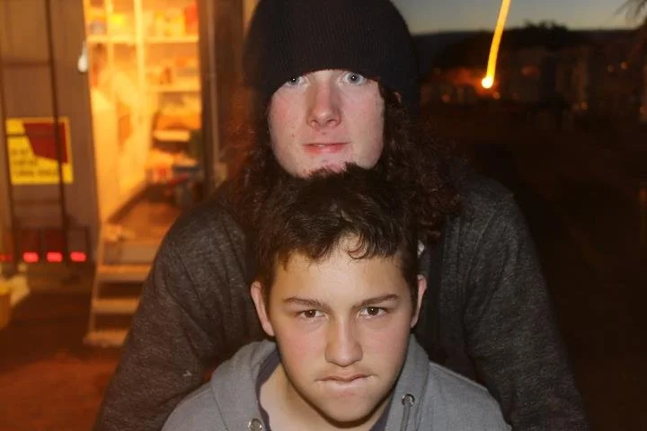 Two young men, one with long curly hair and a black beanie, and the other with short dark hair, are outdoors at nighttime. The person with curly hair is standing behind the person with short hair. They appear to be at a train station or similar locat