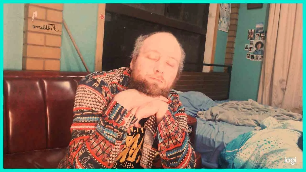 A man with a beard and closed eyes sitting on a bed with his hands clasped under his chin, wearing a colorful patterned jacket.